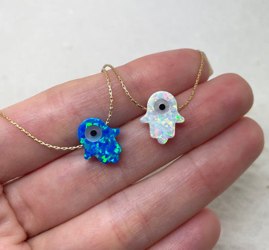 Two Opal Hamsa Evil Eye necklaces laid on a hand, one with a blue opal and the other with a white opal, set against a neutral background.