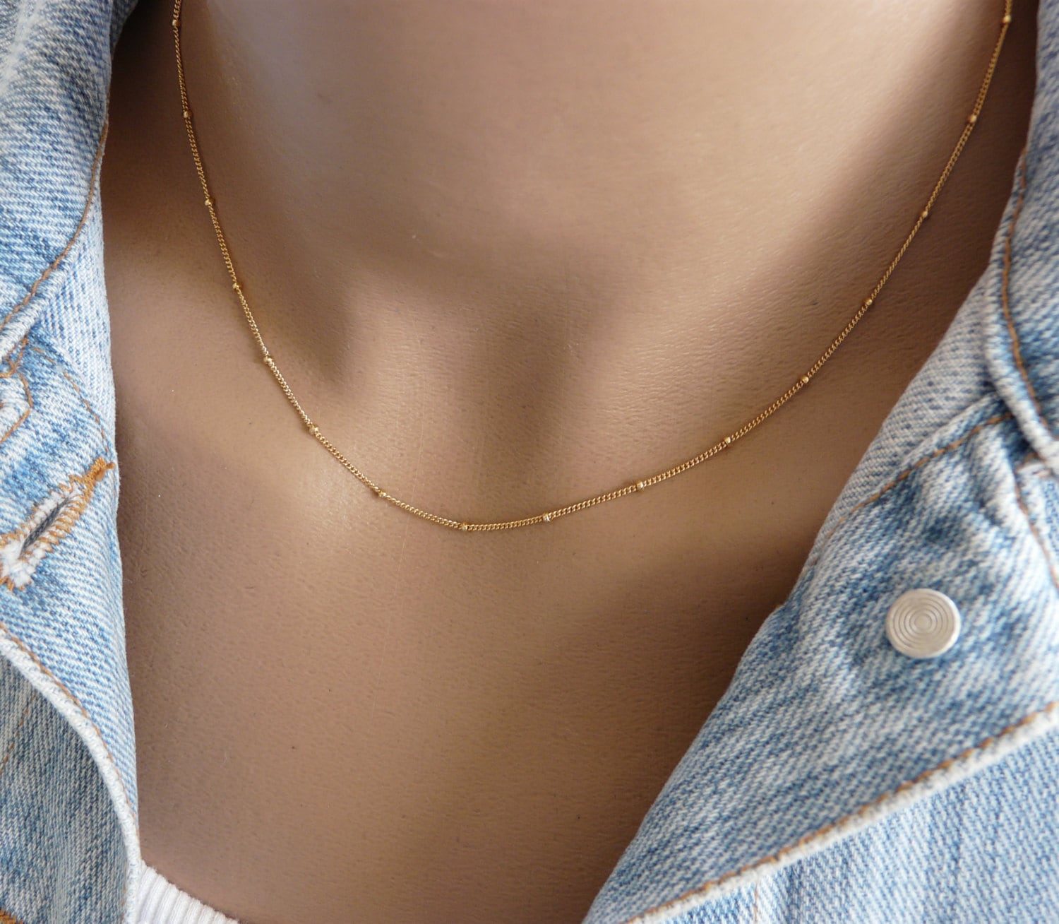A gold-filled satellite chain necklace with a beaded design, displayed on a person wearing a denim jacket.