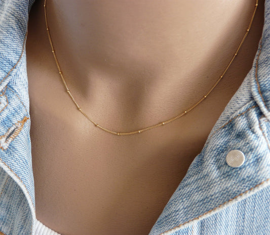 A gold-filled satellite chain necklace with a beaded design, displayed on a person wearing a denim jacket.