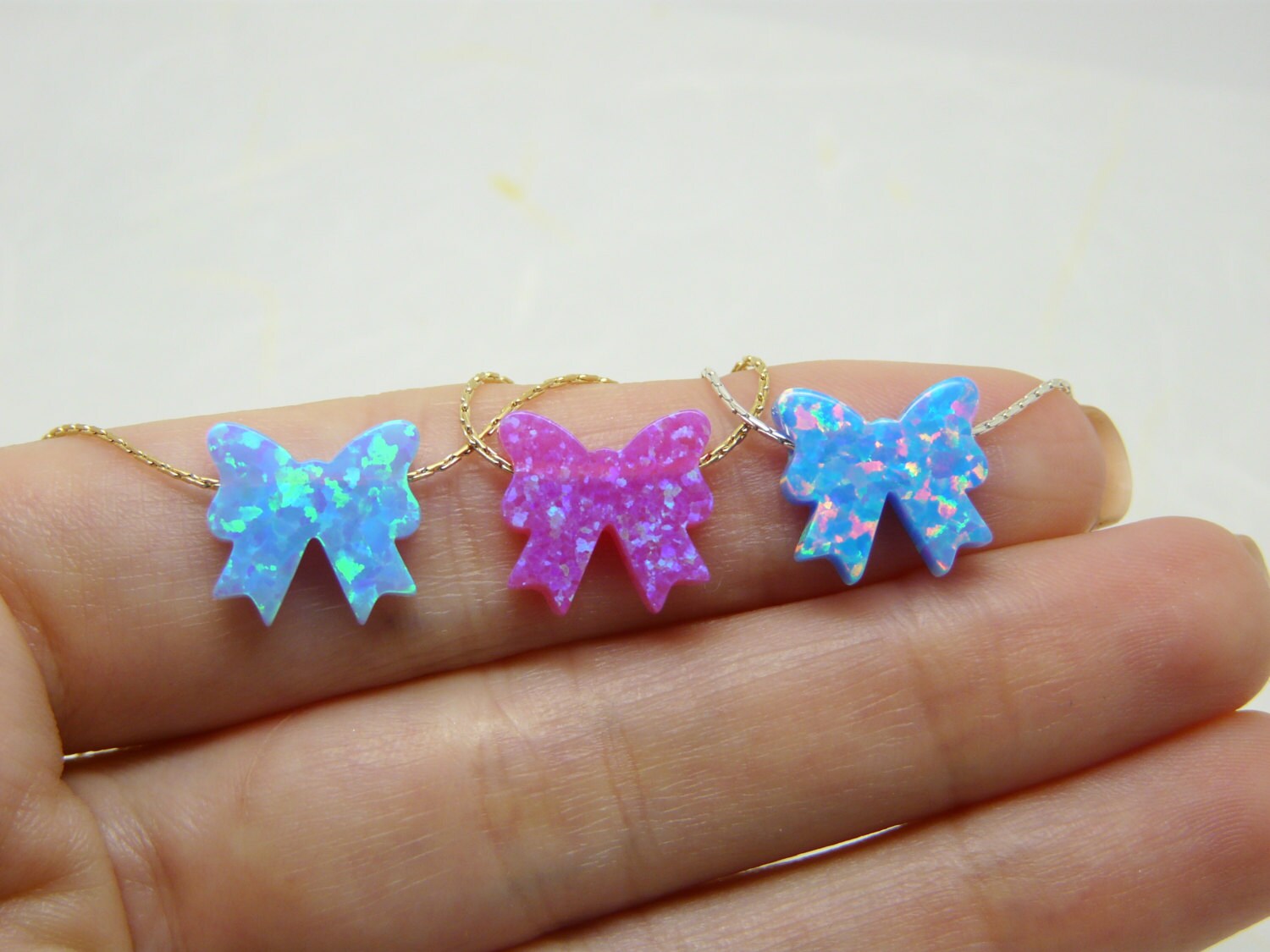A hand holding three bow-shaped necklaces, with the top bow in pink, the middle in white, and the bottom in blue, set against a white background.