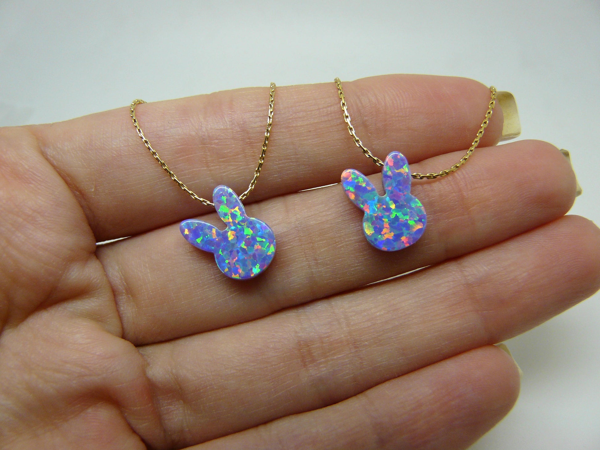Two opal bunny necklaces, one in gold and one in silver color, displayed on a person's hand.