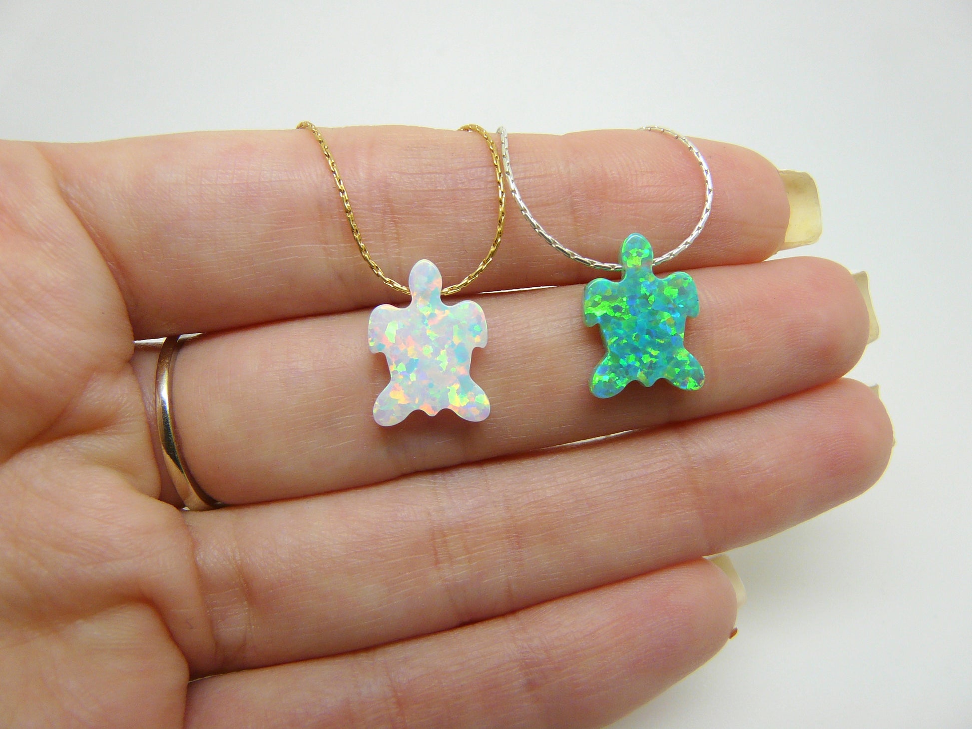 Two opal turtle pendants on gold chains, held in a person's hand. One pendant is white with iridescent qualities, and the other is green with blue undertones.