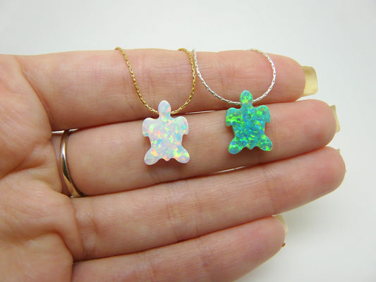 Two opal turtle pendants on gold chains, held in a person's hand. One pendant is white with iridescent qualities, and the other is green with blue undertones.