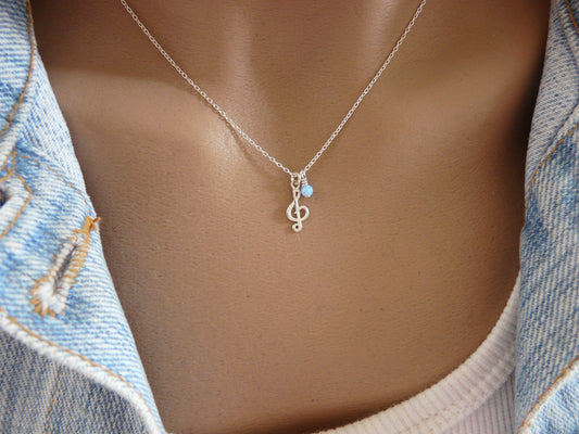 A silver treble clef charm necklace with a small opal bead, displayed on a person wearing a denim jacket.