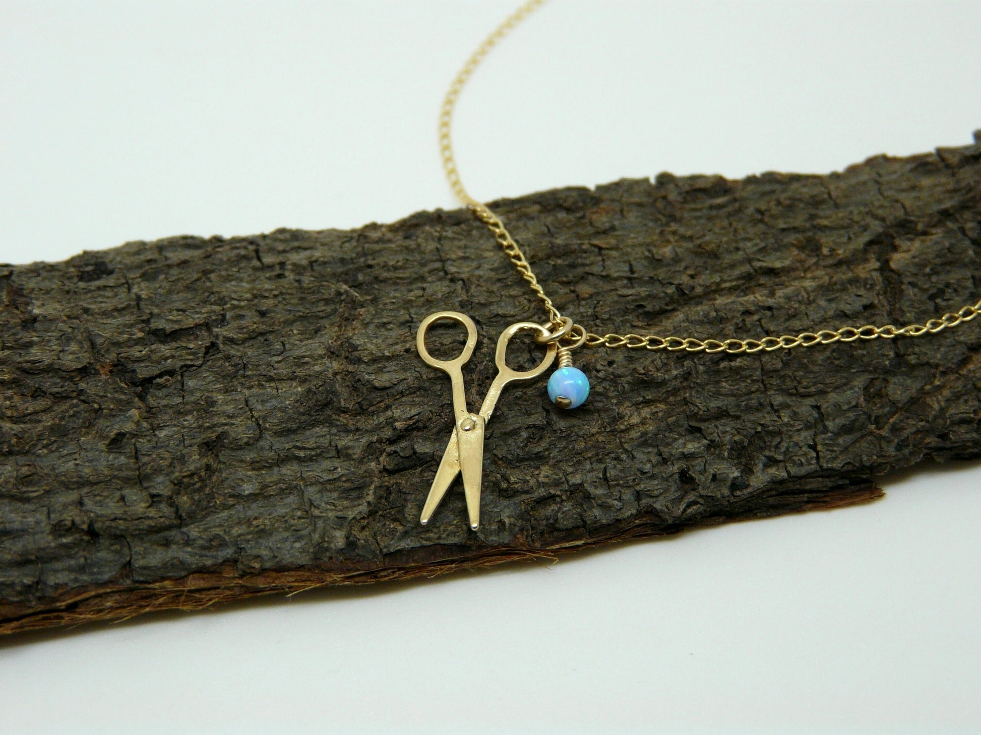 A gold-filled necklace with a scissors charm and a small blue opal bead on a chain, displayed on a wooden branch.