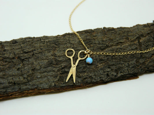A gold-filled necklace with a scissors charm and a small blue opal bead on a chain, displayed on a wooden branch.