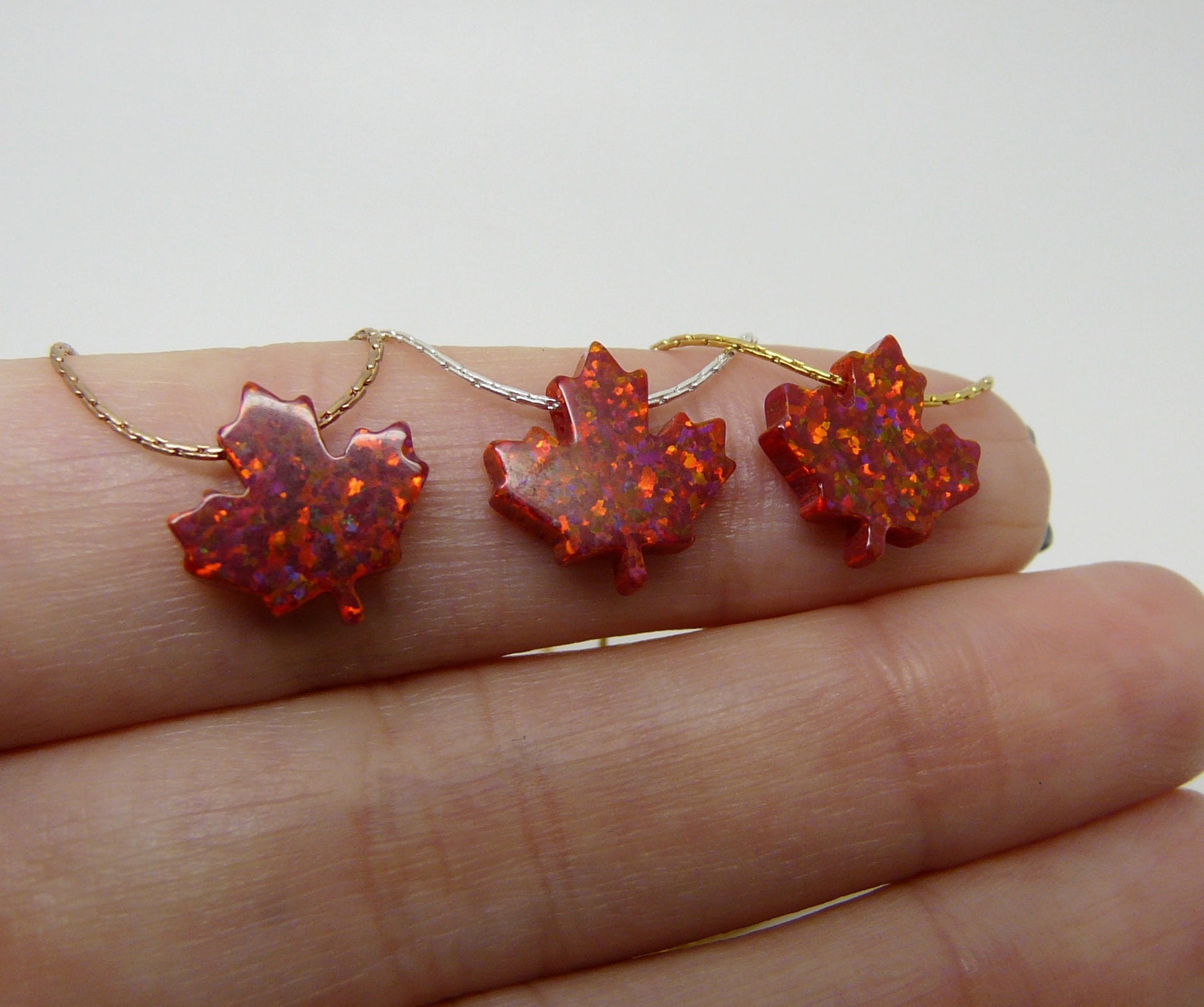 Three opal maple leaf pendants on sterling silver chains, displayed on a person's hand.