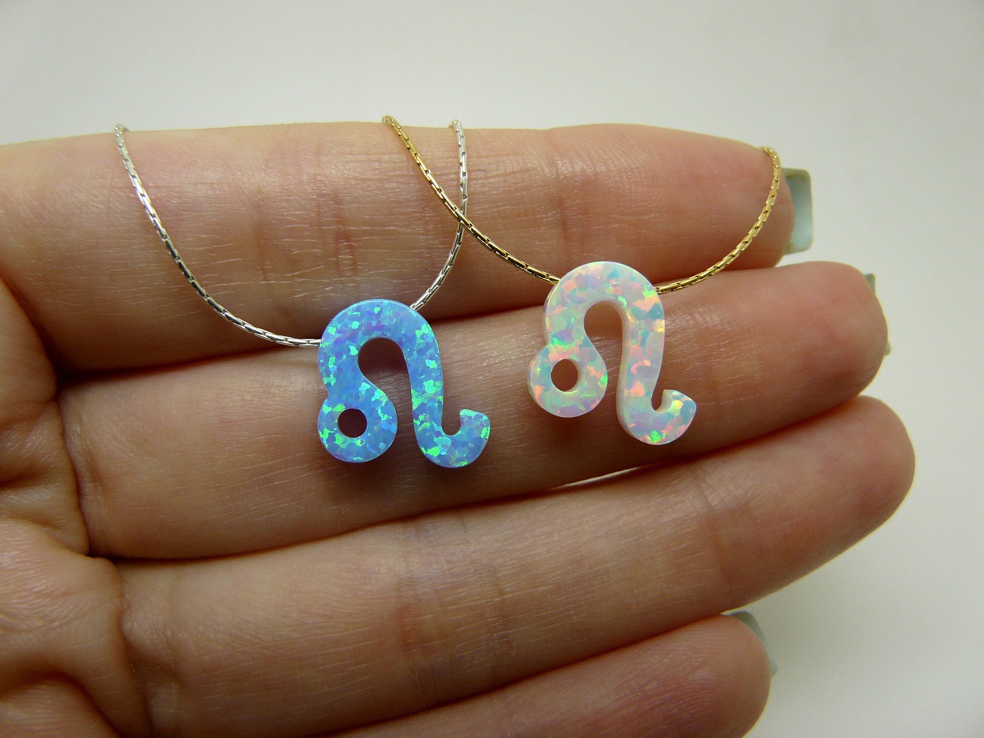 Two opal Leo zodiac pendant necklaces resting on a hand, one with a gold chain and the other with a silver chain.
