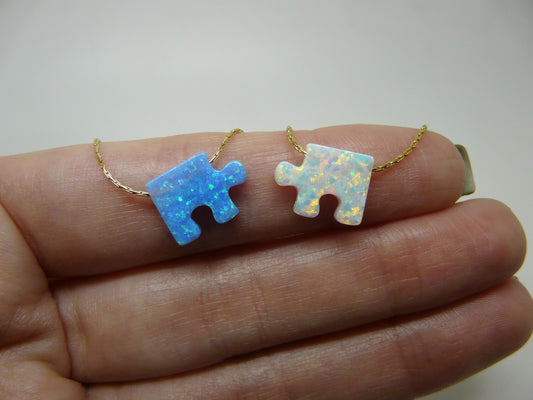 Two opal puzzle piece pendants on gold chains, held in a person's hand.