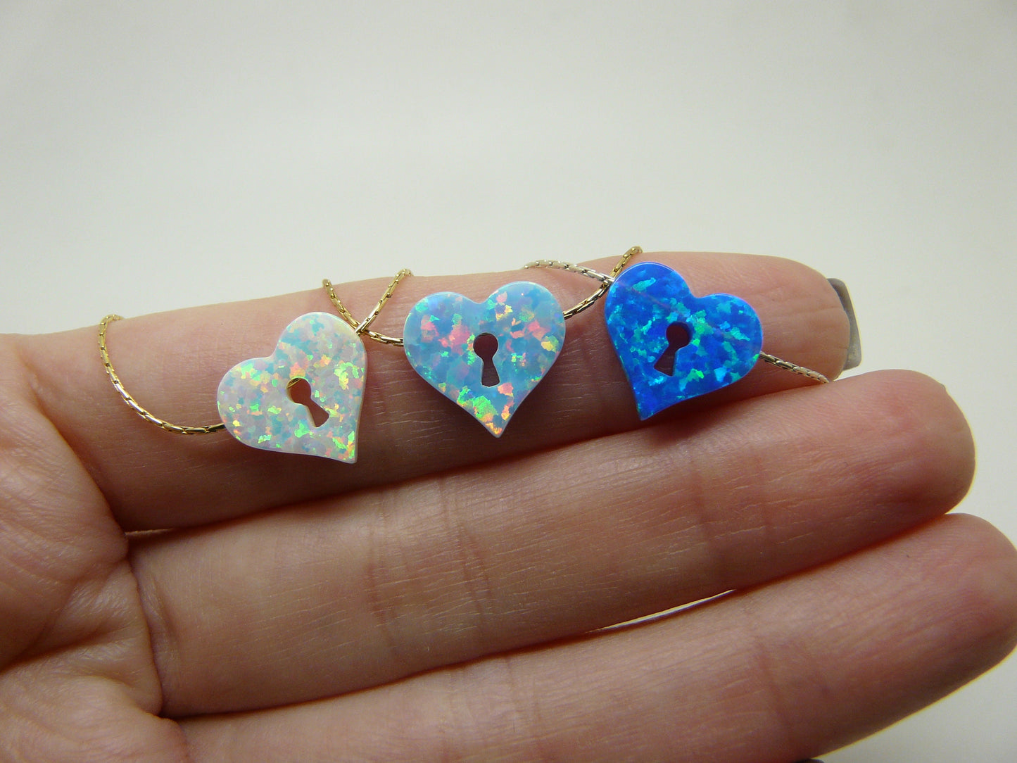 A hand holding three opal heart keyhole necklaces with varying colors.
