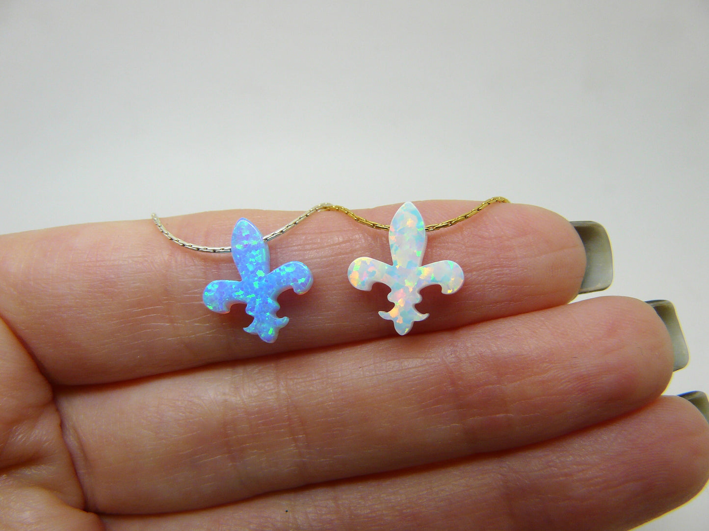 Two opal Fleur de Lis pendants on gold chains, held in a person's hand.