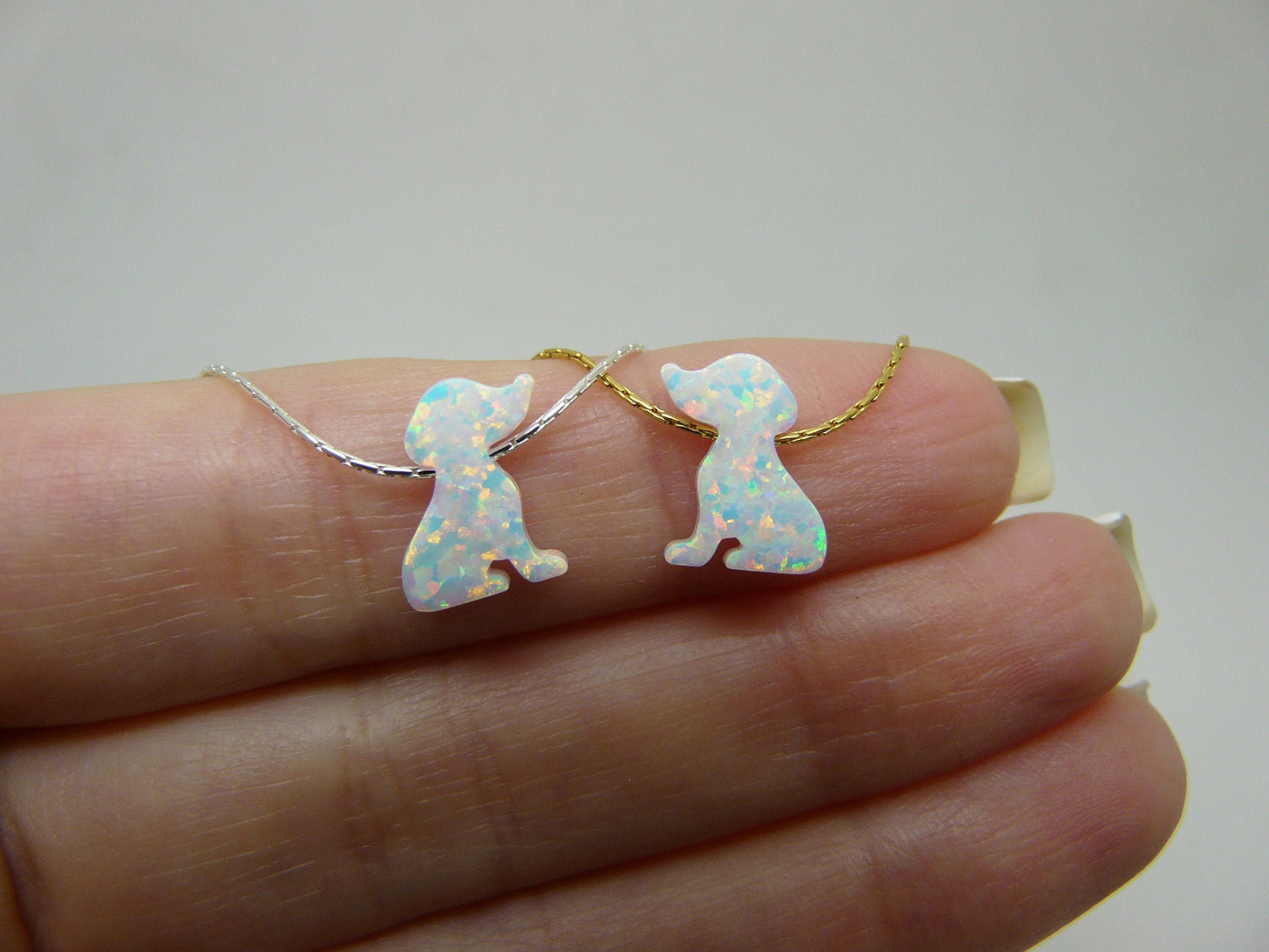 A hand holding two opal dog-shaped pendants on a necklace, one in silver and one in gold filled, against a white background.