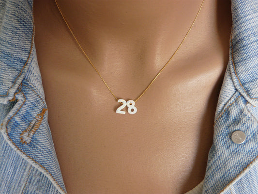 A mannequin wearing a gold number necklace with the number 28 on a white charm, displayed on a person wearing a denim jacket.