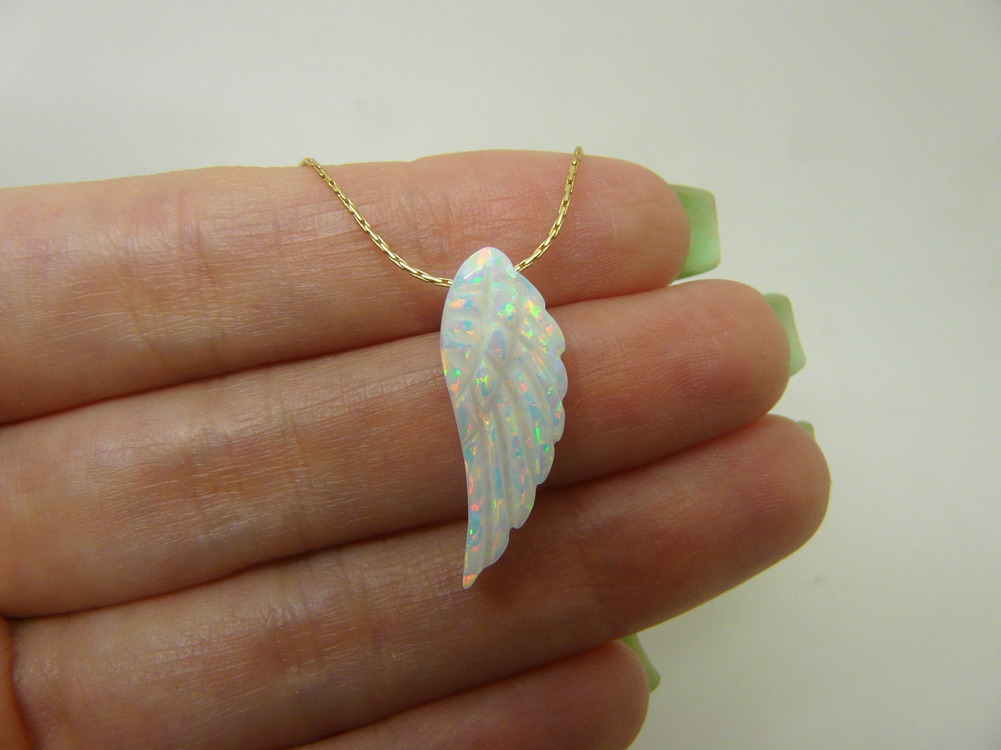 A hand holding a delicate necklace with a carved opal angel wing pendant attached to a gold-filled or sterling silver chain.