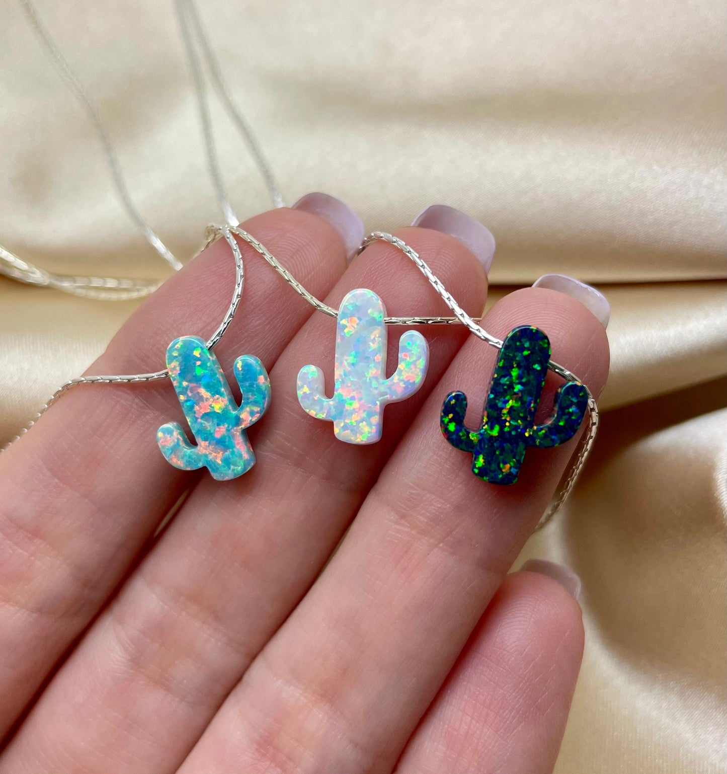 A hand holding three opal cactus necklaces with pendants in blue, white, and black colors, suspended on silver chains.