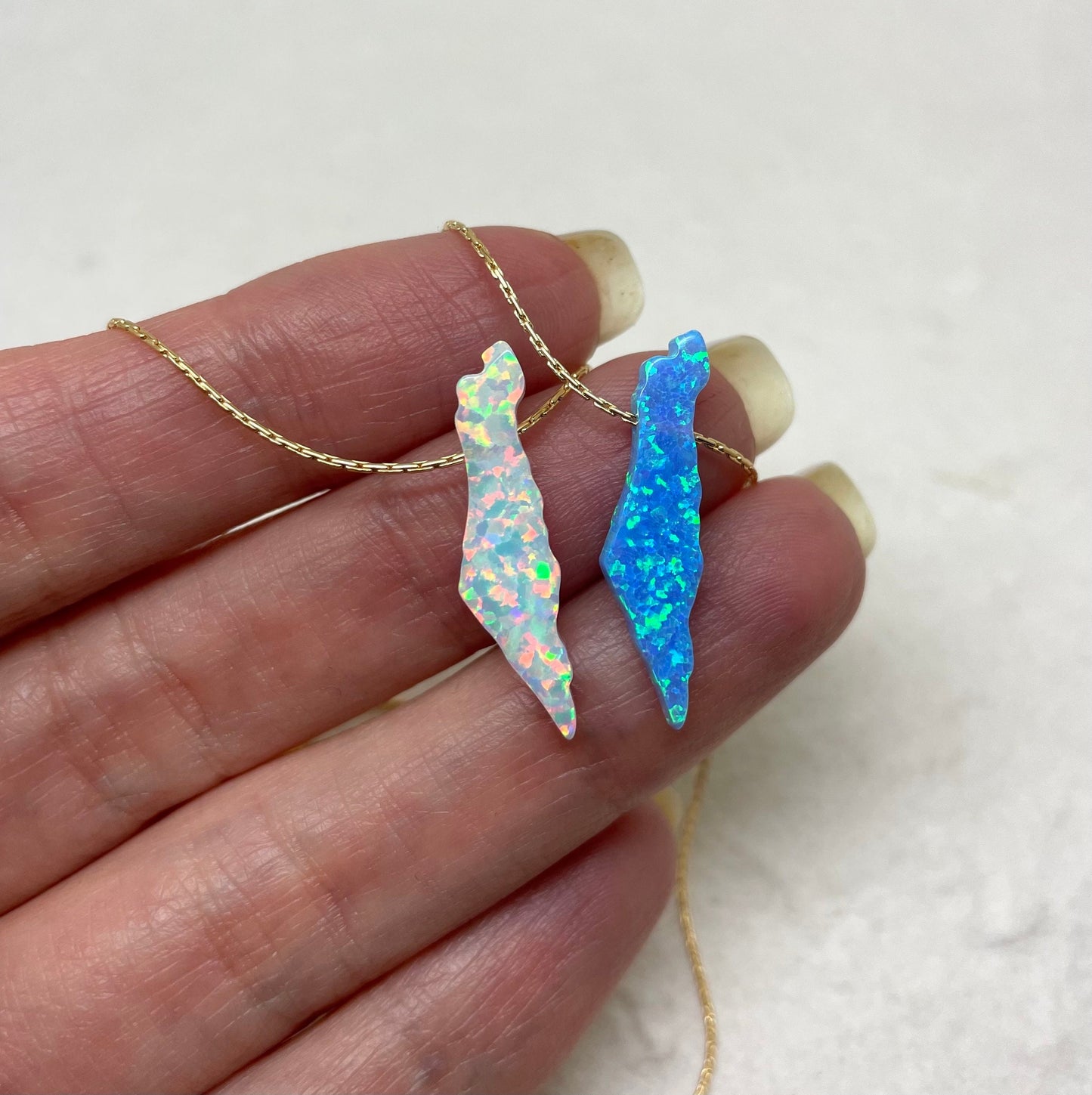 Two Israel map pendants on necklaces, one with a pink opal finish and the other with a blue opal finish, held in a person's hand.