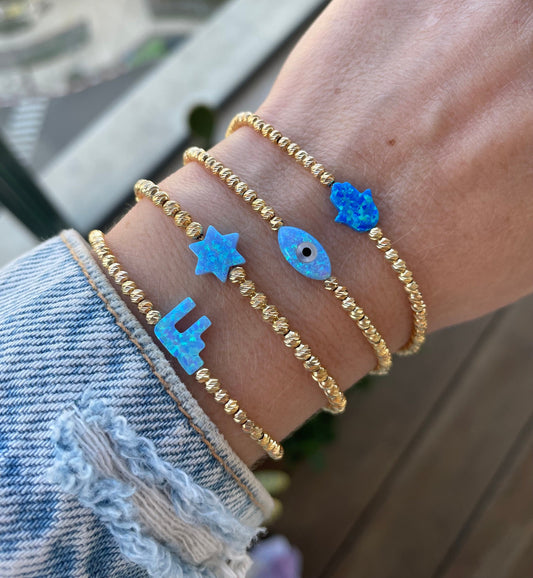 A close-up image of a person's wrist wearing a gold-colored bracelet with blue opal charms in the shapes of the Hamsa Hand, Star of David, Chai, and Evil Eye.