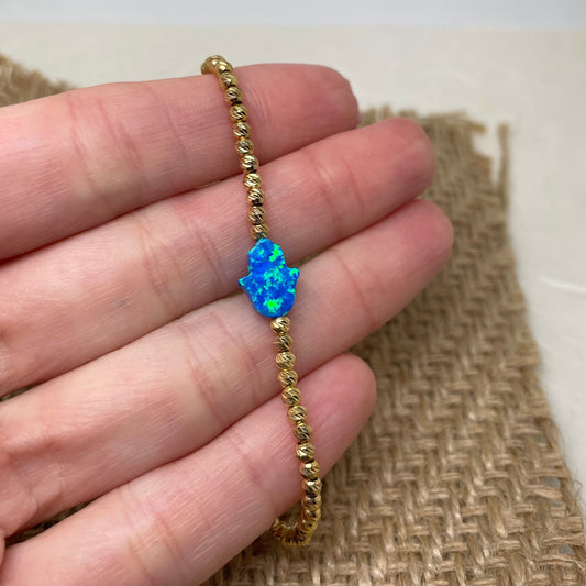 A hand holding a gold-filled bracelet with a blue opal Hamsa charm and small gold beads.