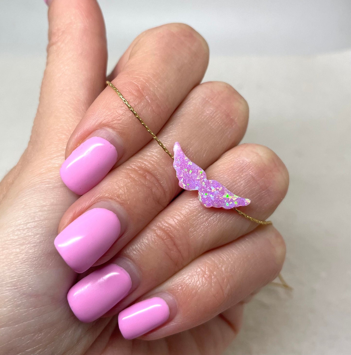 A hand with pink nail polish holding a pink opal angel wing pendant necklace on a gold chain.