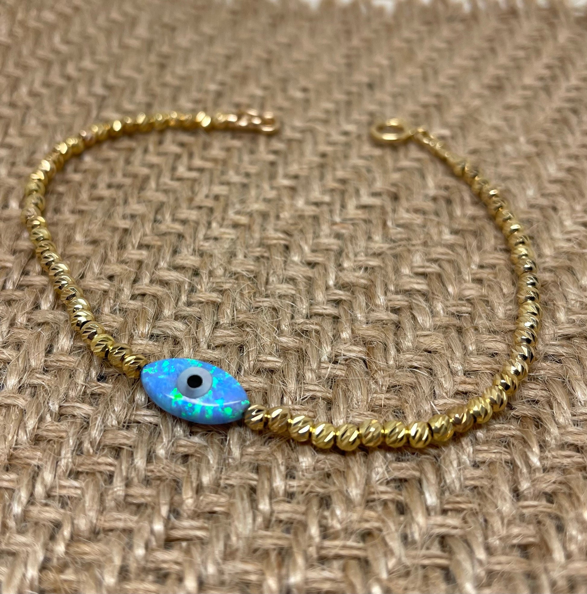 A gold-colored bracelet featuring tiny beads and an opal evil eye charm, displayed on a textured background.