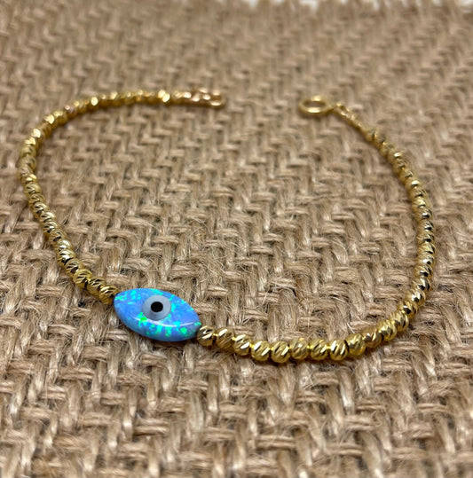A gold-colored bracelet featuring tiny beads and an opal evil eye charm, displayed on a textured background.