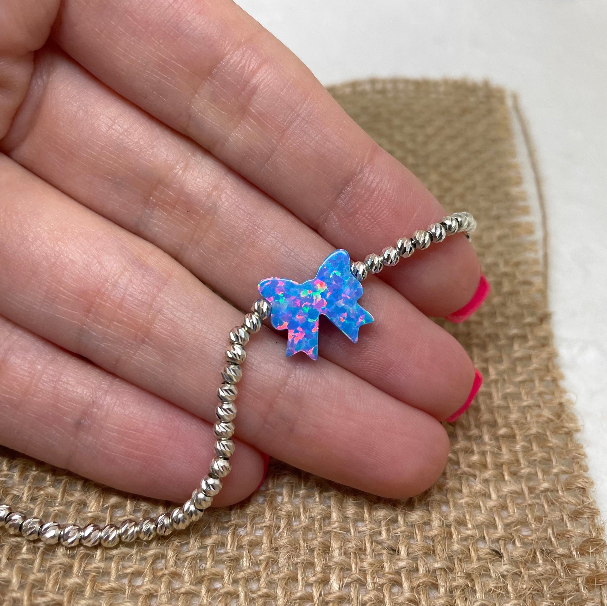 A hand holding a bracelet featuring a blue opal bow charm on a silver chain, with the bow charm facing upwards.