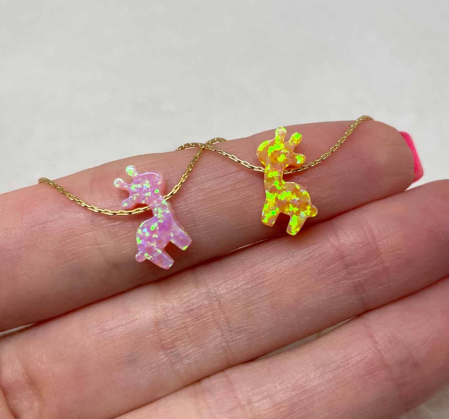 Two giraffe-shaped pendants on necklaces, one pink and one yellow, placed on a person's finger.