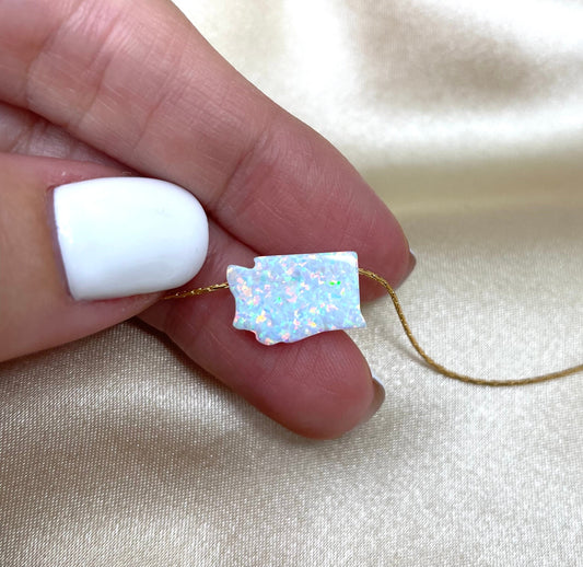 A hand holding a Washington-shaped opal pendant necklace on a gold or yellow-colored chain.