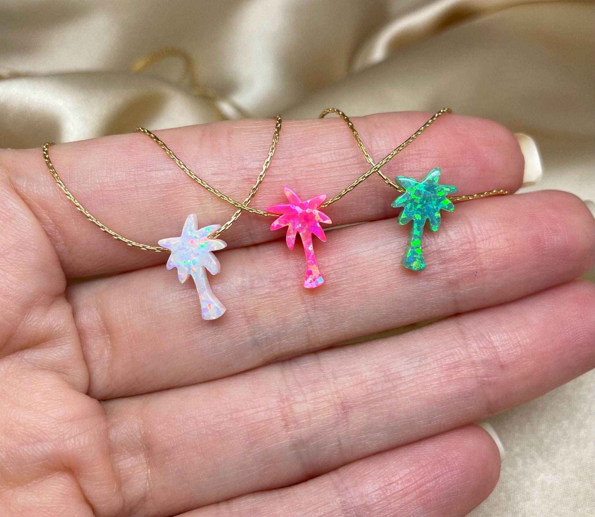 Three palm tree pendants on necklaces, in pink, white, and green colors, held in a hand.