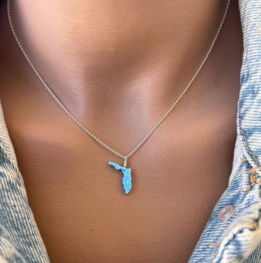 A person wearing a denim jacket showcasing a necklace with a Florida state-shaped opal pendant.