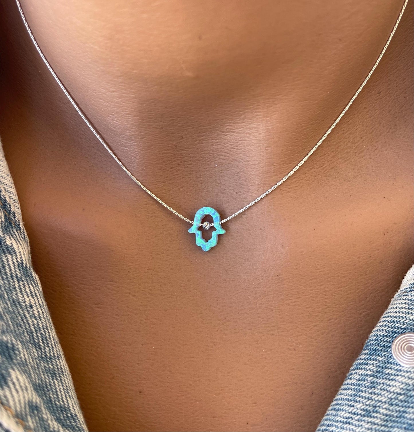 A person wearing a blue opal Hamsa necklace with a silver chain, displayed against a casual clothing backdrop.