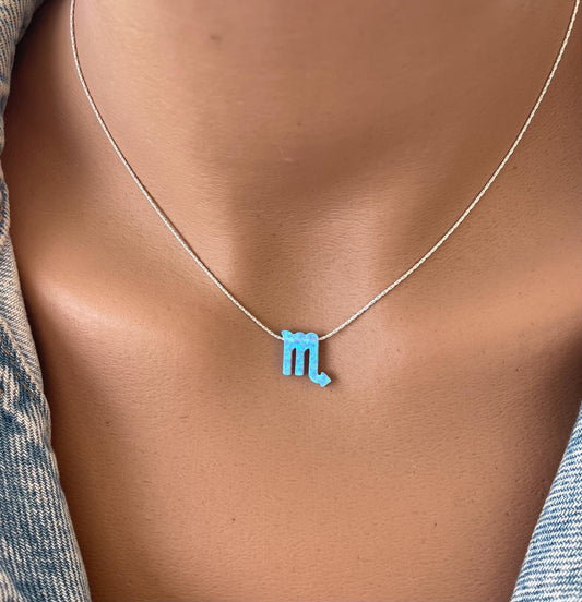 A person wearing a necklace with a small, blue opal pendant shaped like the Scorpio zodiac symbol, displayed on a mannequin.