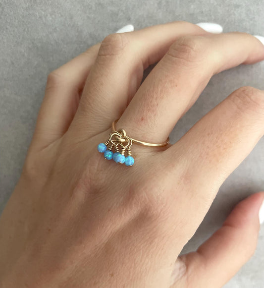 A hand wearing a gold-filled band ring with tiny blue opal beads dangling on the end.