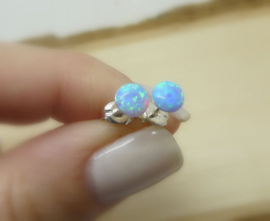 A person holding a pair of tiny blue opal stud earrings, showing the blue opal stones reflecting light.
