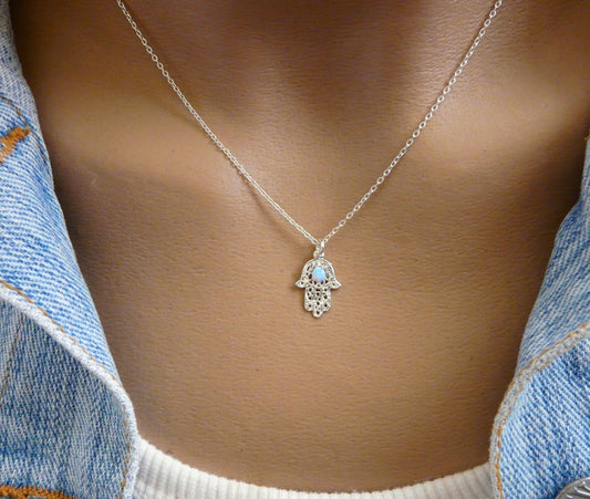 A sterling silver Hamsa pendant necklace with an opal bead accent, worn by a person with a denim jacket in the background.