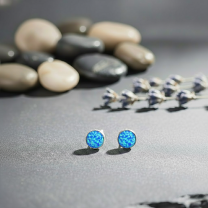 Blue opal stud earrings on a gray surface with pebbles and flowers in the background