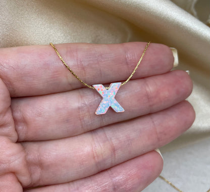 Gold necklace with a pink 'x' pendant held in a hand against a beige fabric background