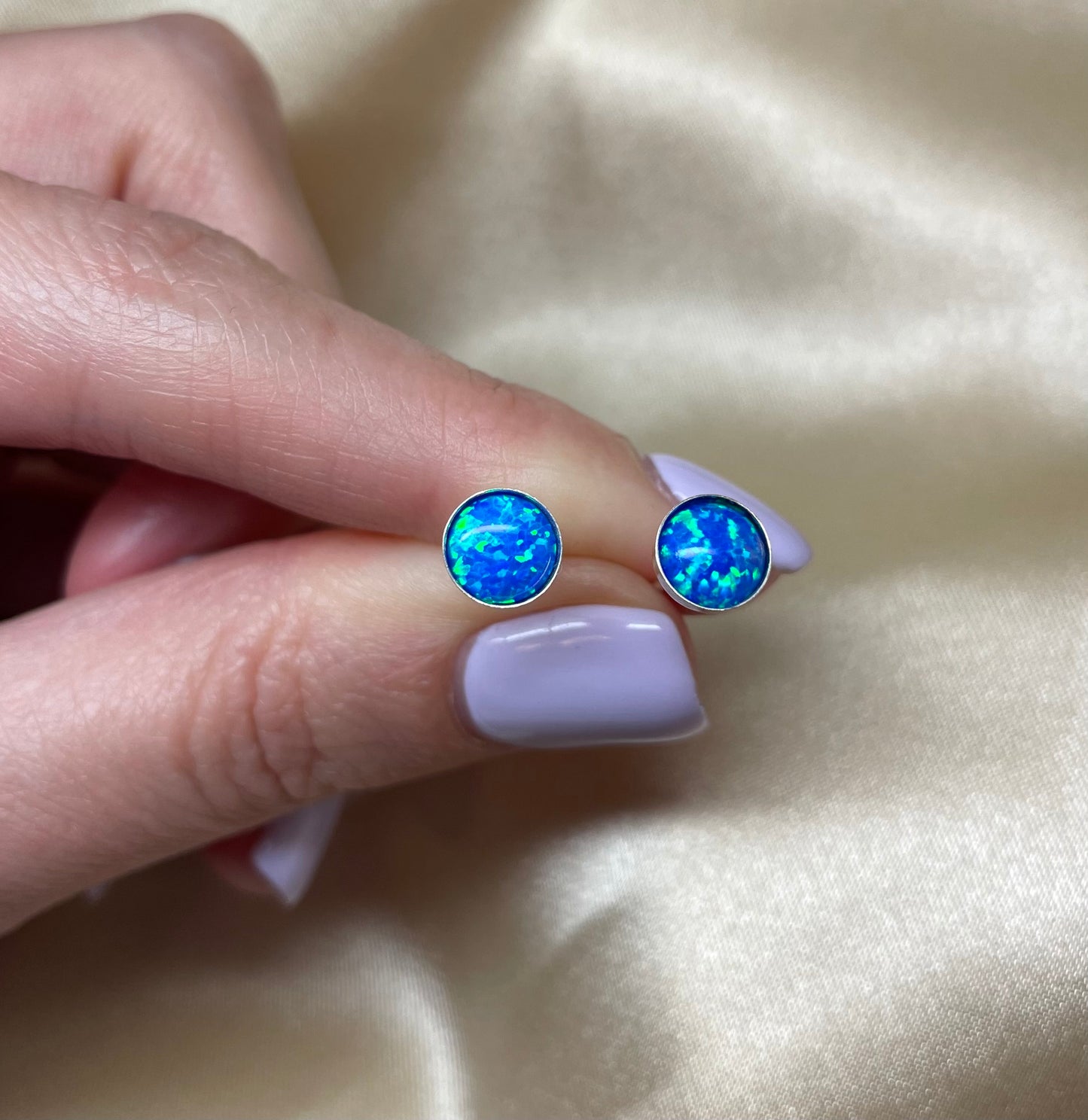 Blue opal stud earrings held between fingers on a beige fabric background