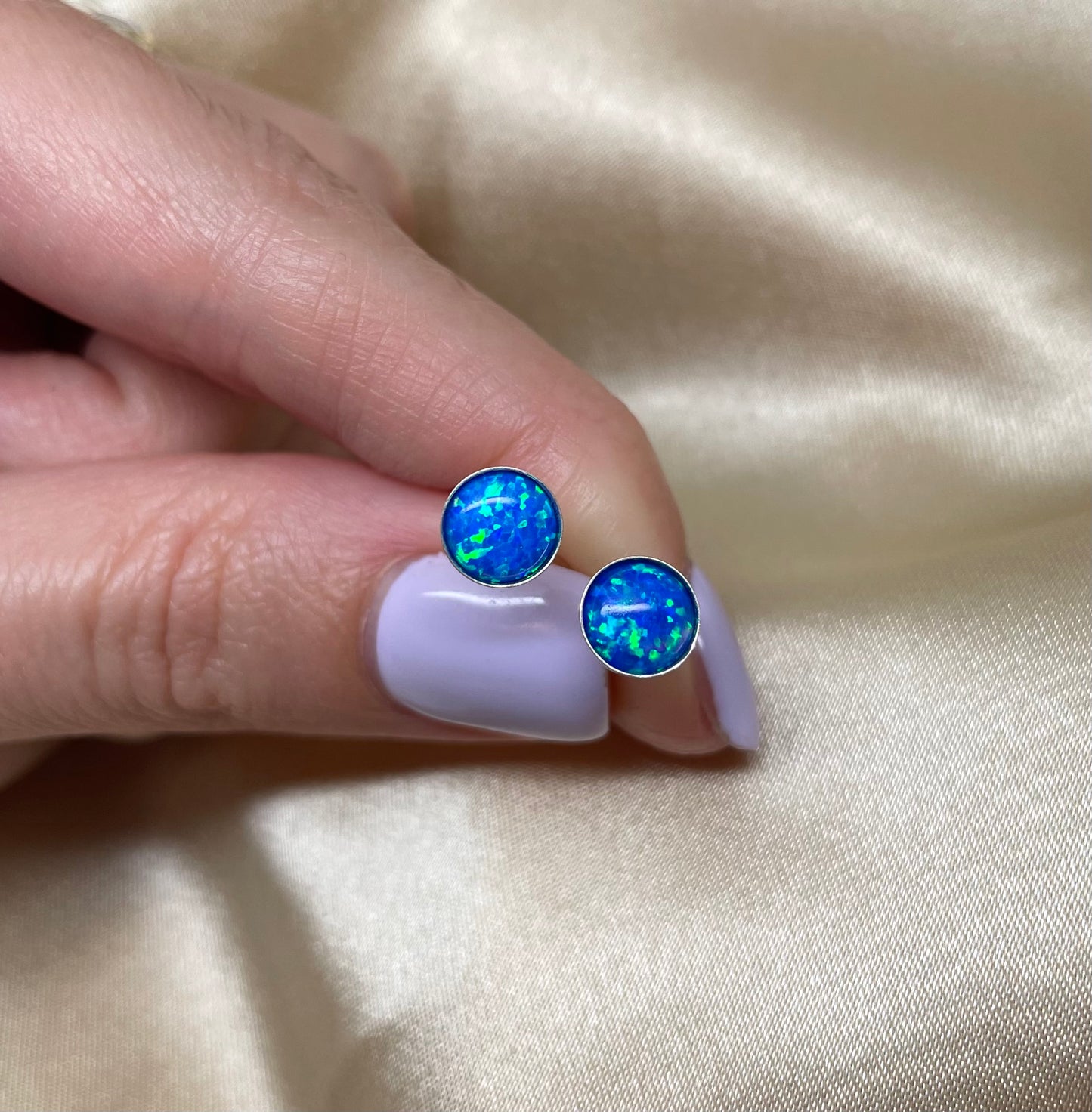Blue opal stud earrings held between fingers on a beige fabric background