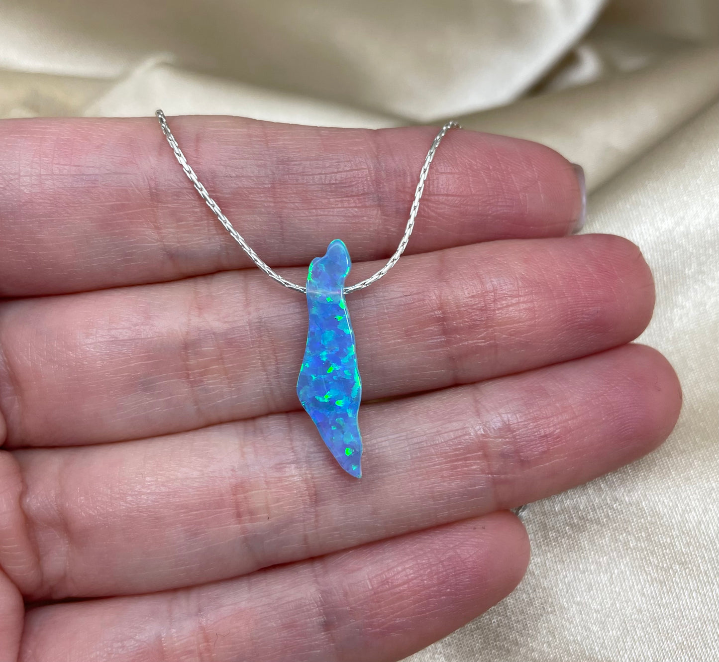 Blue opal pendant on a silver chain held in a hand against a beige fabric background